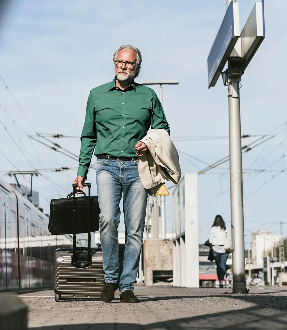 Ein Fahrgast mit Koffer und Jacke über den Arm gehängt, geht bei sommerlichem Wetter über einen Bahnsteig (themenunterstützendes Bild)